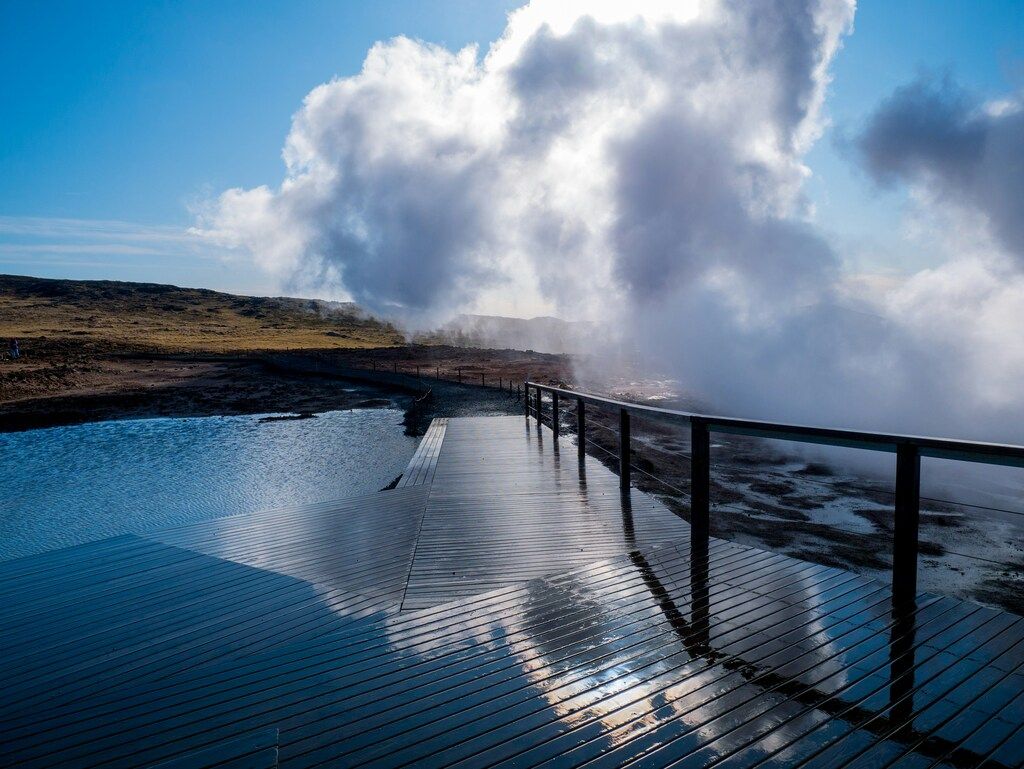 Eine Promenade führt über eine dampfende geothermische Quelle, mit klarem Himmel und karger Landschaft im Hintergrund.