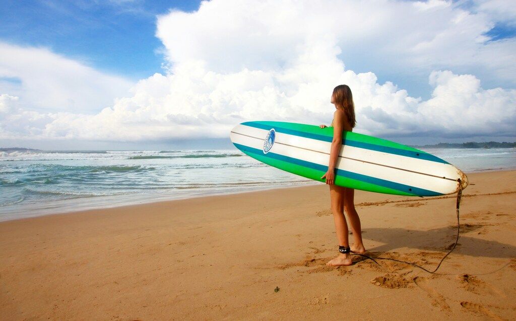 Frau steht am Meer mit weißem, blauem und grünem Surfbrett unter blauem Himmel