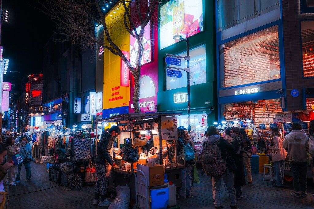 Belebte Einkaufsstraße bei Nacht mit bunten Leuchtreklamen und Marktständen.