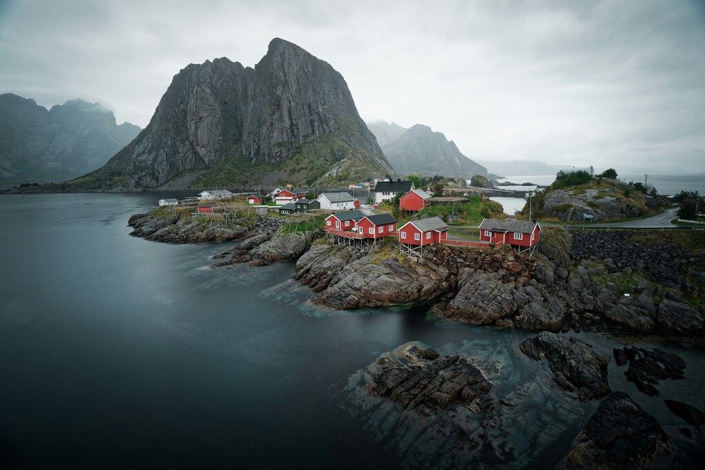 Lofoten Sehenswürdigkeiten: Rote Fischerhütten auf Felsen vor einer dramatischen Bergkulisse in einer ruhigen Meereslandschaft.