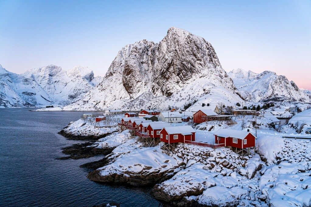 Schneebedeckte Fjordlandschaft mit roten Hütten vor einem großen Berg in Norwegen.