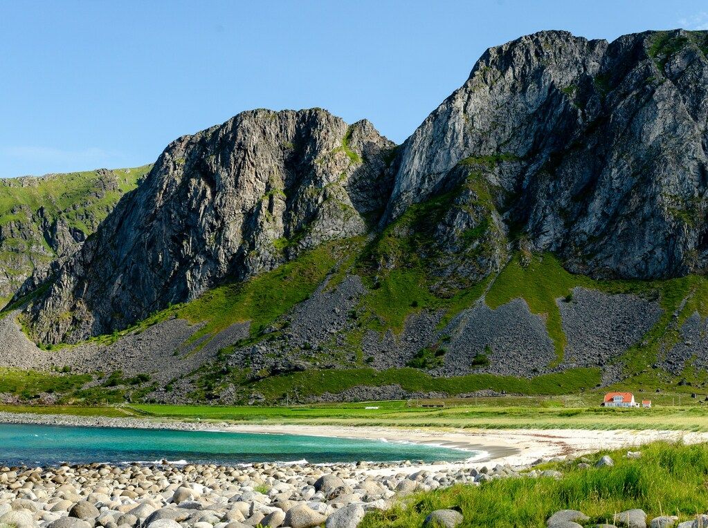 Ein idyllischer Strand mit türkisfarbenem Wasser, umgeben von grünen Wiesen und steilen Felsklippen unter klarem blauem Himmel.