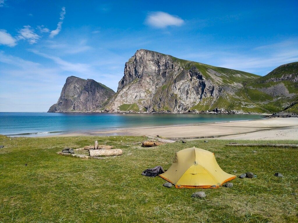 Ein Zelt auf einer grünen Wiese vor einem ruhigen Strand mit türkisfarbenem Wasser und beeindruckenden Klippen im Hintergrund