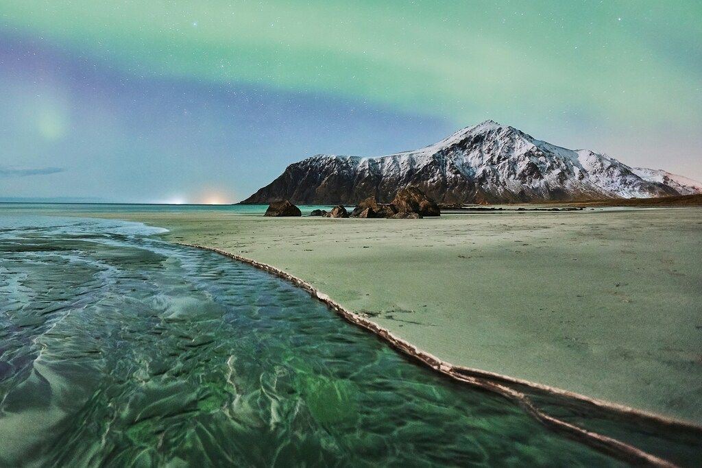 Grüne Nordlichter über einem Strand mit Bergen im Hintergrund.