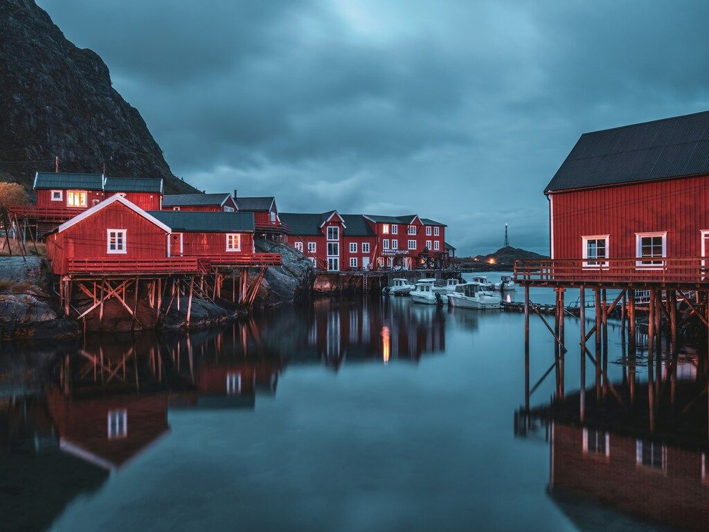 Lofoten Sehenswürdigkeiten: Beleuchtete rote Fischerhütten spiegeln sich in ruhigem Wasser unter einem bewölkten Himmel.