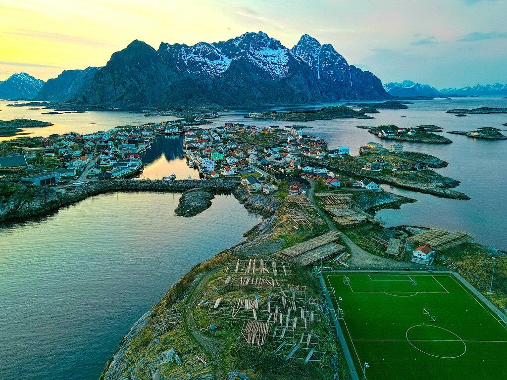 Ein Dorf mit bunten Häusern und einem Fußballplatz, umgeben von Wasser und schneebedeckten Bergen.