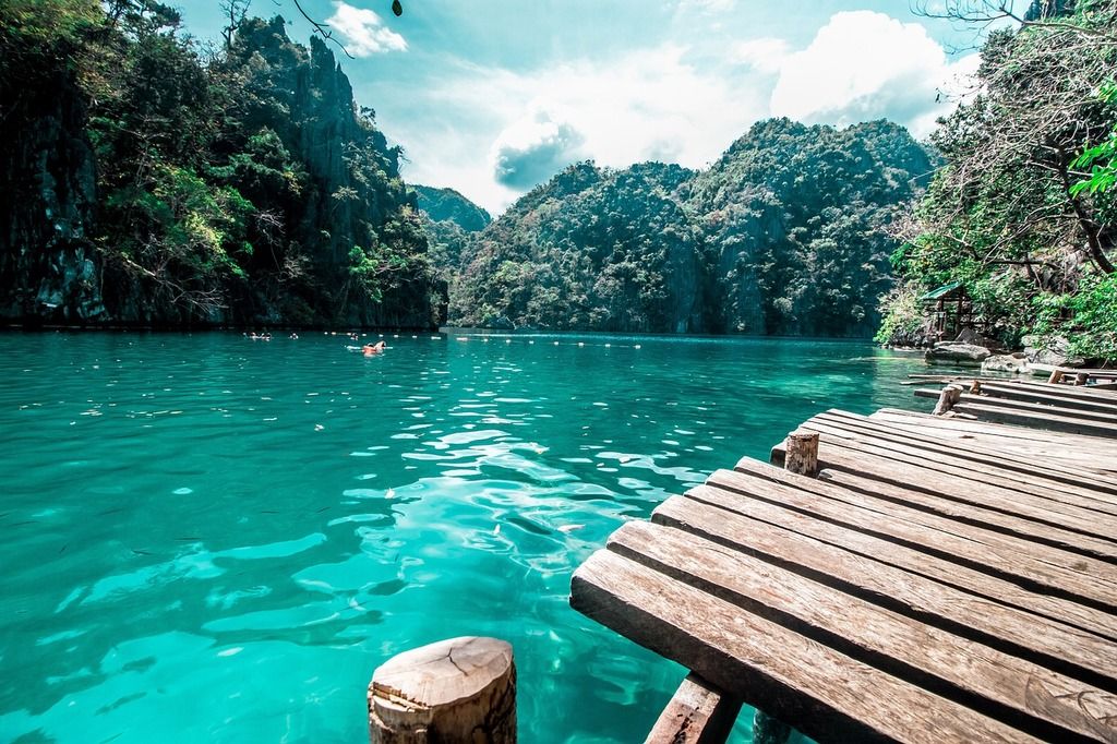 Holzsteg am Coron-See mit türkisblauem wasser und steilen felsen im hintergrund.
