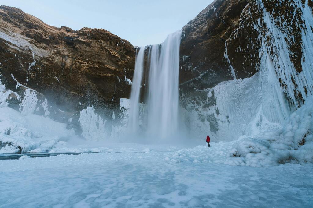 Der gefrorene Skógafoss-Wasserfall im Winter mit einer roten Figur in der Ferne.
