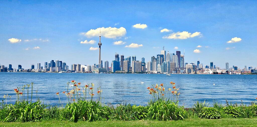 Panoramablick auf die Skyline von Toronto mit dem CN Tower im Hintergrund, gesehen von einem grünen Ufer.
