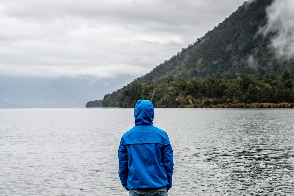 Eine Person in einer blauen Jacke blickt auf einen ruhigen See mit bewaldeten Bergen im Hintergrund.