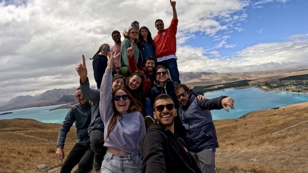 Eine Gruppe von Reisenden lacht und feiert auf einem Hügel mit Blick auf einen türkisblauen See während einer Weroad-Gruppenreise in Neuseeland.