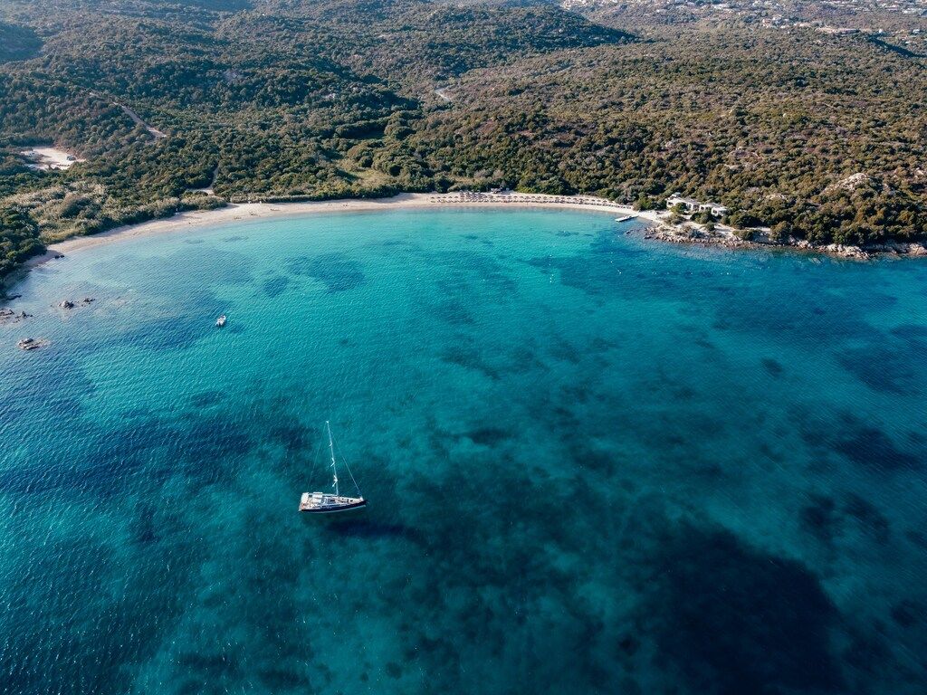 Luftaufnahme einer Bucht in der Costa Smeralda auf Sardinien mit Boot und bewaldeten Hügeln.