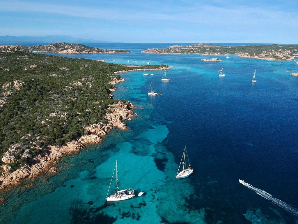 Boote in klarem blauem Wasser des Archipels La Maddalena auf Sardinien vor felsiger Küste.