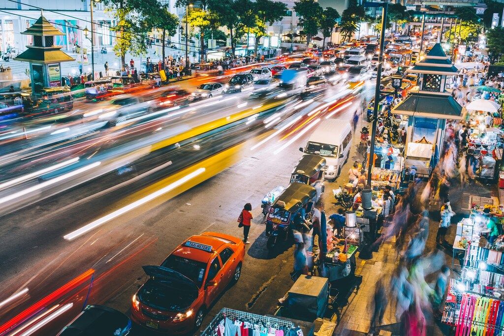 Nachtaufnahme einer belebten Straße in Bangkok mit Tuk-Tuks, Marktständen und Lichtspuren.