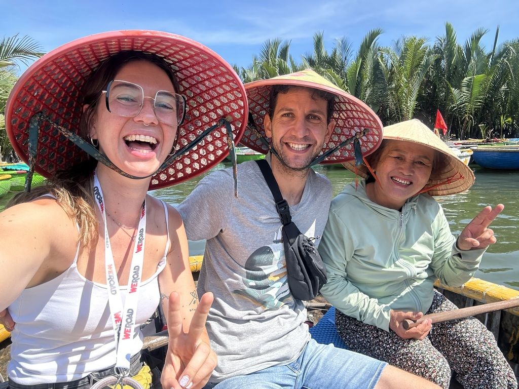Drei lachende Menschen mit traditionellen vietnamesischen Hüten machen ein Selfie auf einem Boot zwischen Palmen in Vietnam.