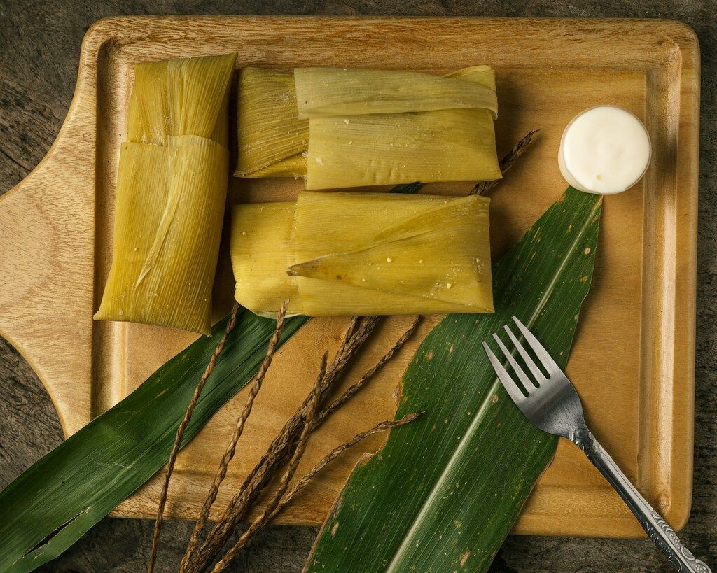 Traditionelle Tamales in Maisblättern serviert mit Sahne auf einem Holzbrett.