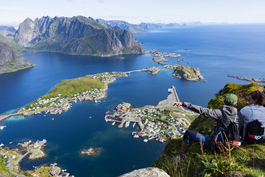 Zwei Reisende genießen die spektakuläre Aussicht vom Reinebringen auf das Insel-Dorf Reine und das blaue Meer.