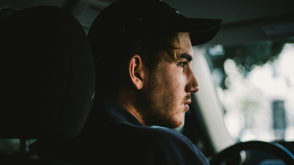 Ein junger Mann mit einer schwarzen Kappe, im Profil in einem Auto sitzend.