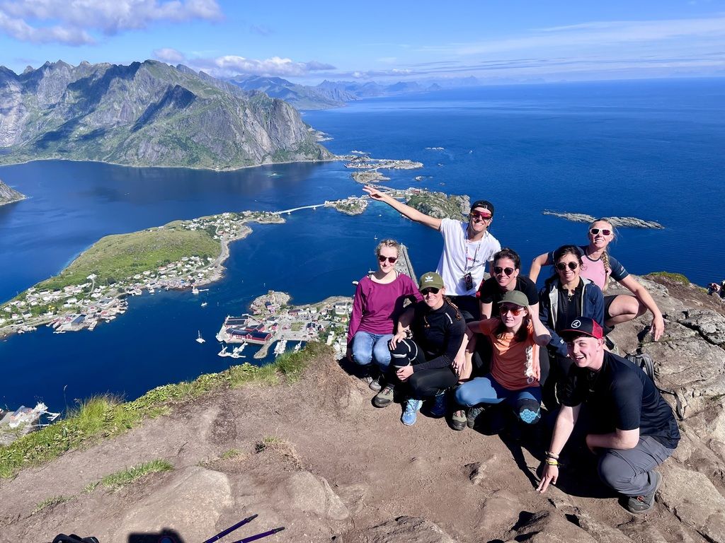 WeRoad-Reisegruppe posiert auf dem Gipfel mit atemberaubender Aussicht über Reine und das tiefblaue Nordmeer.