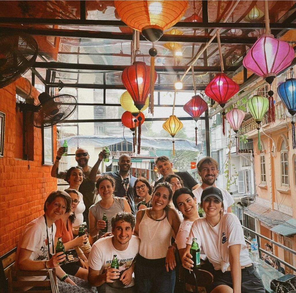 Gruppe von Freunden auf einem Balkon in Vietnam umgeben von bunten Laternen, toastend mit Bier.