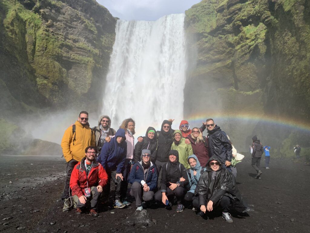 WeRoad-Gruppe macht Gruppenfoto vor dem Skógafoss-Wasserfall in Island mit Regenbogen.