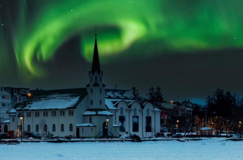 Die Nordlichter (Aurora Borealis) tanzen grün über einer kleinen, weißen Kirche in Reykjavik im winterlichen Schnee. Die Nordlichter (Aurora Borealis) tanzen grün über einer kleinen, weißen Kirche in Reykjavik im winterlichen Schnee.