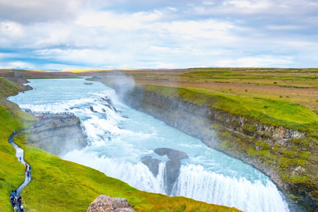 Breiter Wasserfall Gullfoss in Island mit Touristen auf dem Aussichtspfad und grüner Landschaft.