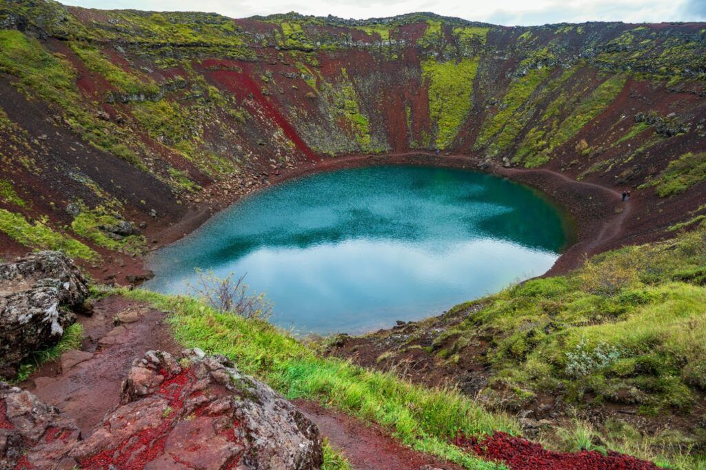 Blick auf den leuchtend blauen Kratersee Kerið, umgeben von roter und grüner Erde in Island.