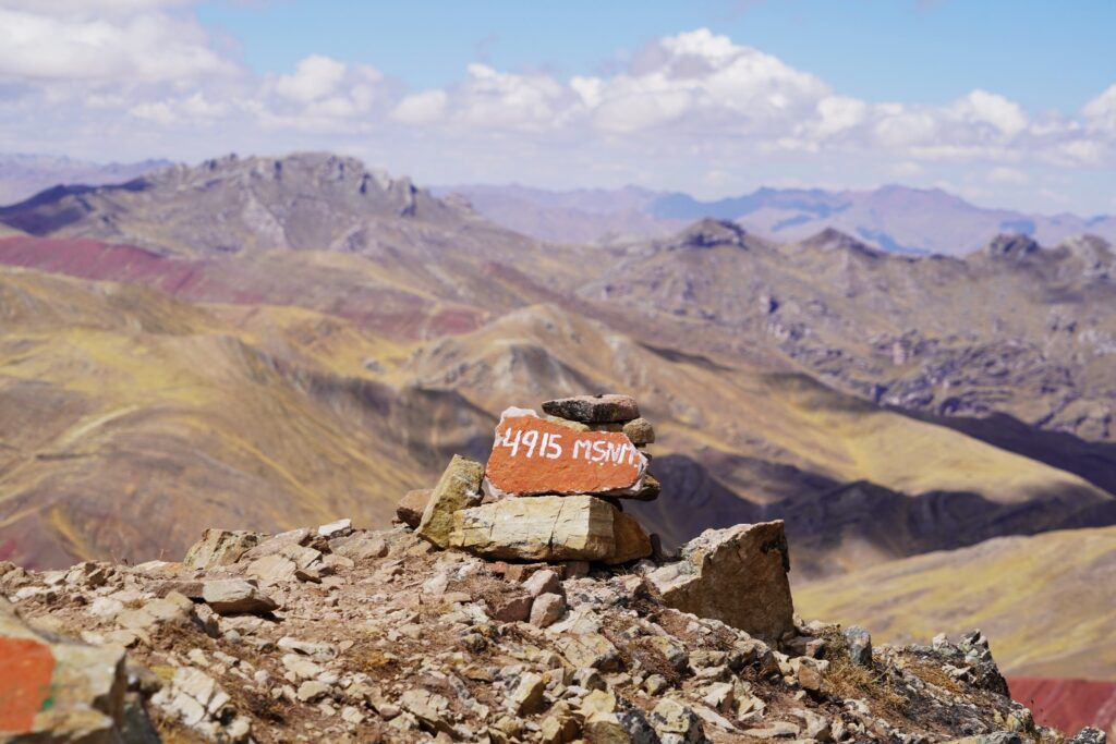 Steinhaufen mit einer roten Tafel „4915 MSNM“ auf dem Gipfel mit Blick auf die farbenfrohen Berge der Anden in Peru.