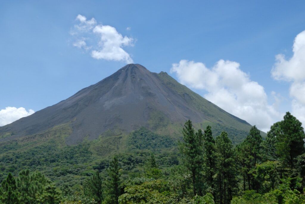 Der beeindruckende Kegel des Vulkans Arenal in Costa Rica unter einem blauen Himmel mit ein paar weißen Wolken, umgeben von dichtem grünem Wald.