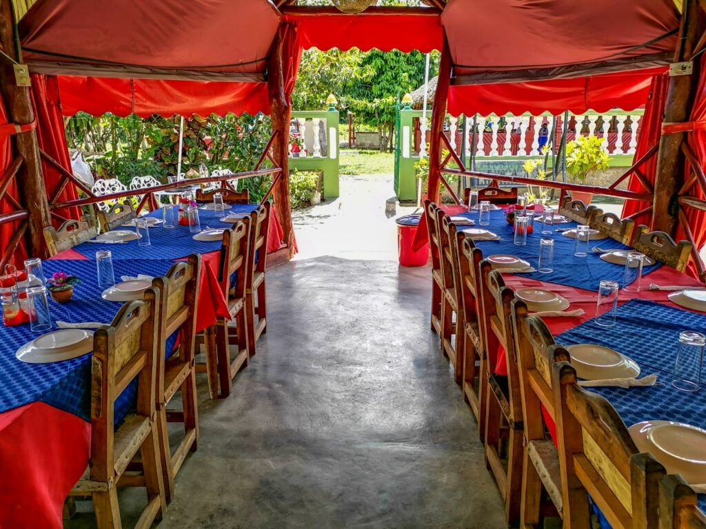 Long wooden dining tables with blue tablecloths set for a group meal in an outdoor covered terrace in Cuba.