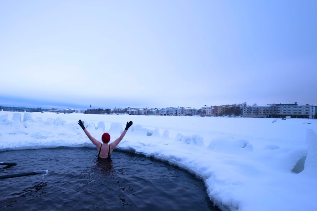 Eine Person mit roter Mütze und schwarzen Handschuhen badet mit erhobenen Armen in einem Eisloch, umgeben von einer dicken Schneedecke und einer winterlichen Stadtkulisse im Hintergrund.