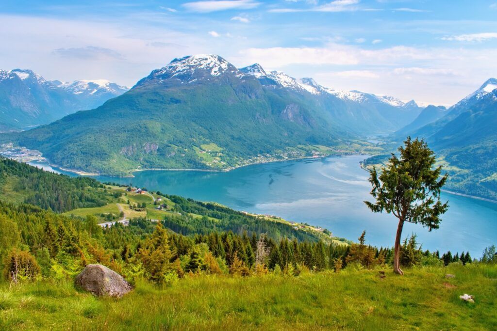 Blick auf den ruhigen, blauen Nordfjord in Norwegen, umgeben von grünen Tälern und schneebedeckten Bergen unter einem sonnigen Himmel.