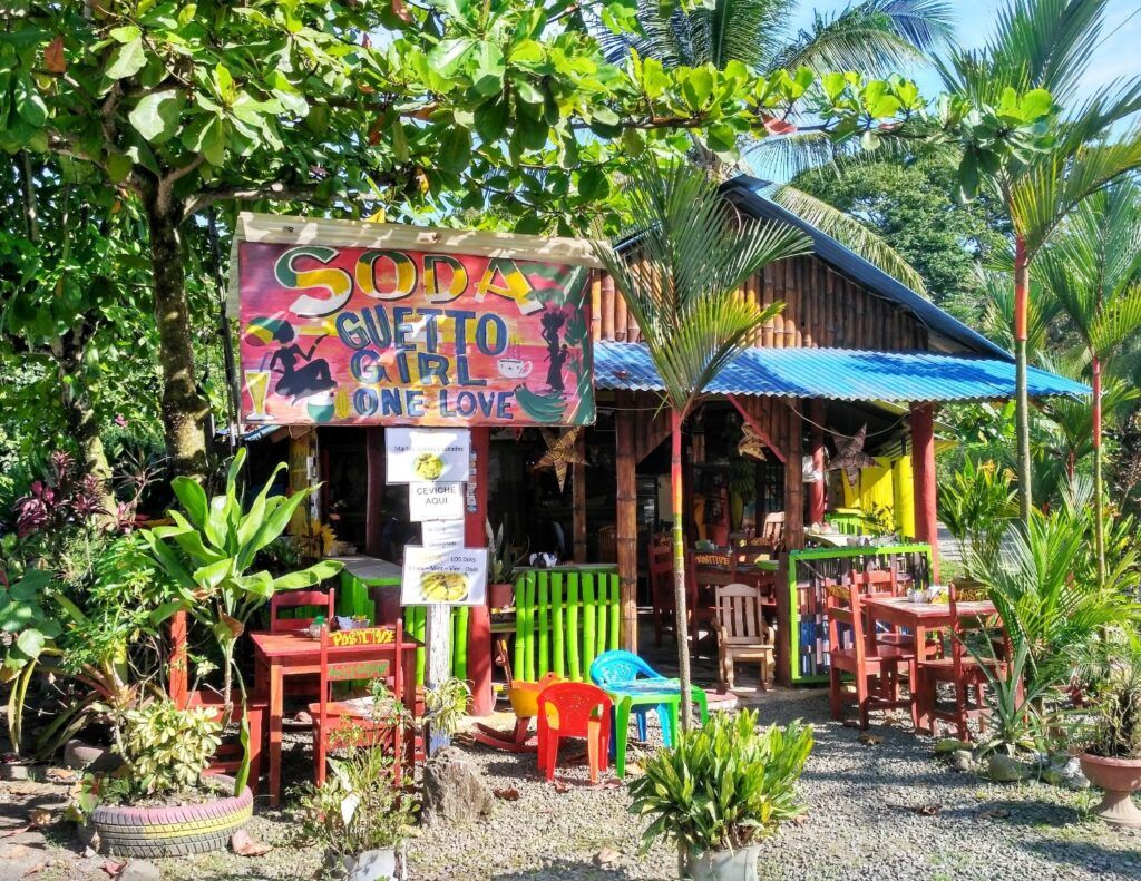Ein farbenfrohes lokales Restaurant namens 'Soda Guetto Girl' in Costa Rica, mit einem handgemalten Schild, roten Holztischen und Stühlen im Freien unter Palmen.