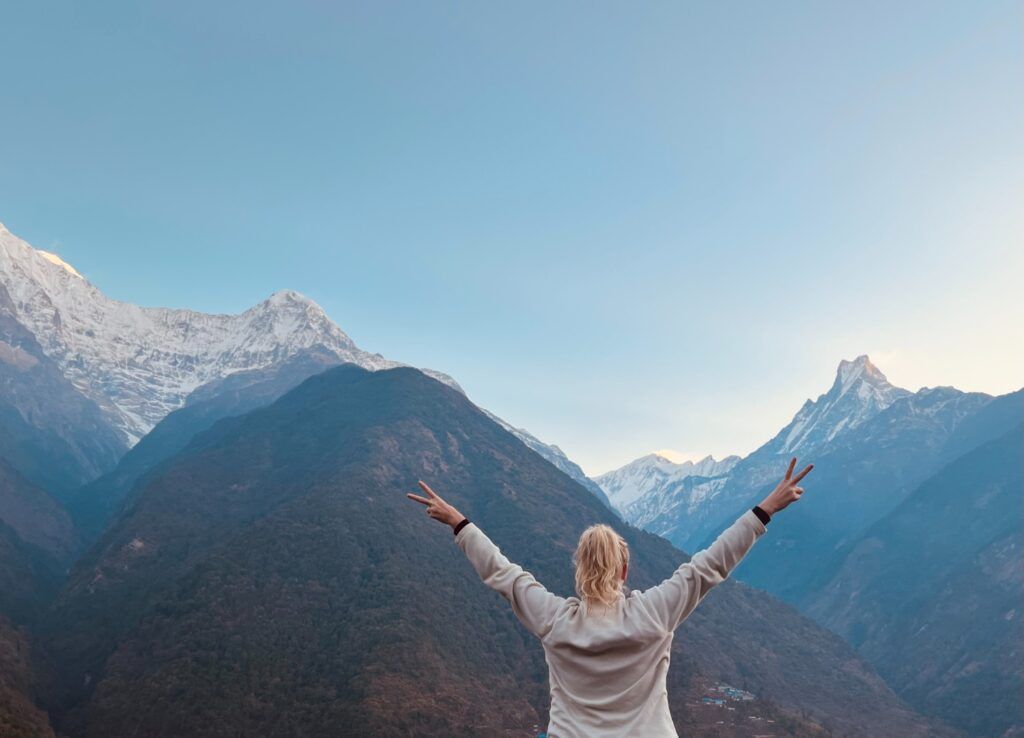 Eine Person steht mit dem Rücken zur Kamera und hebt die Arme im Peace-Zeichen vor den schneebedeckten Gipfeln des Himalayas in Nepal.