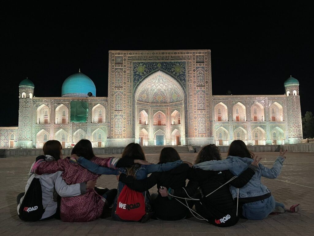 Eine Gruppe von WeRoad-Reisenden sitzt nachts vor der beleuchteten Tilla-Kori-Madrasa in Samarkand, Usbekistan.