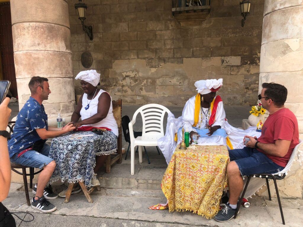 Dos viajeros sentados en mesas al aire libre con dos mujeres locales en ropa blanca tradicional en La Habana.
