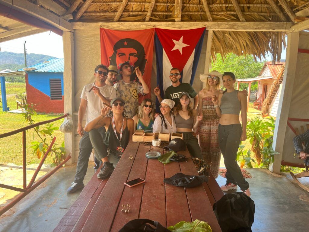 Un grupo de viajeros WeRoad posando juntos bajo una cabaña en Cuba, con la bandera cubana y un retrato del Che Guevara al fondo.
