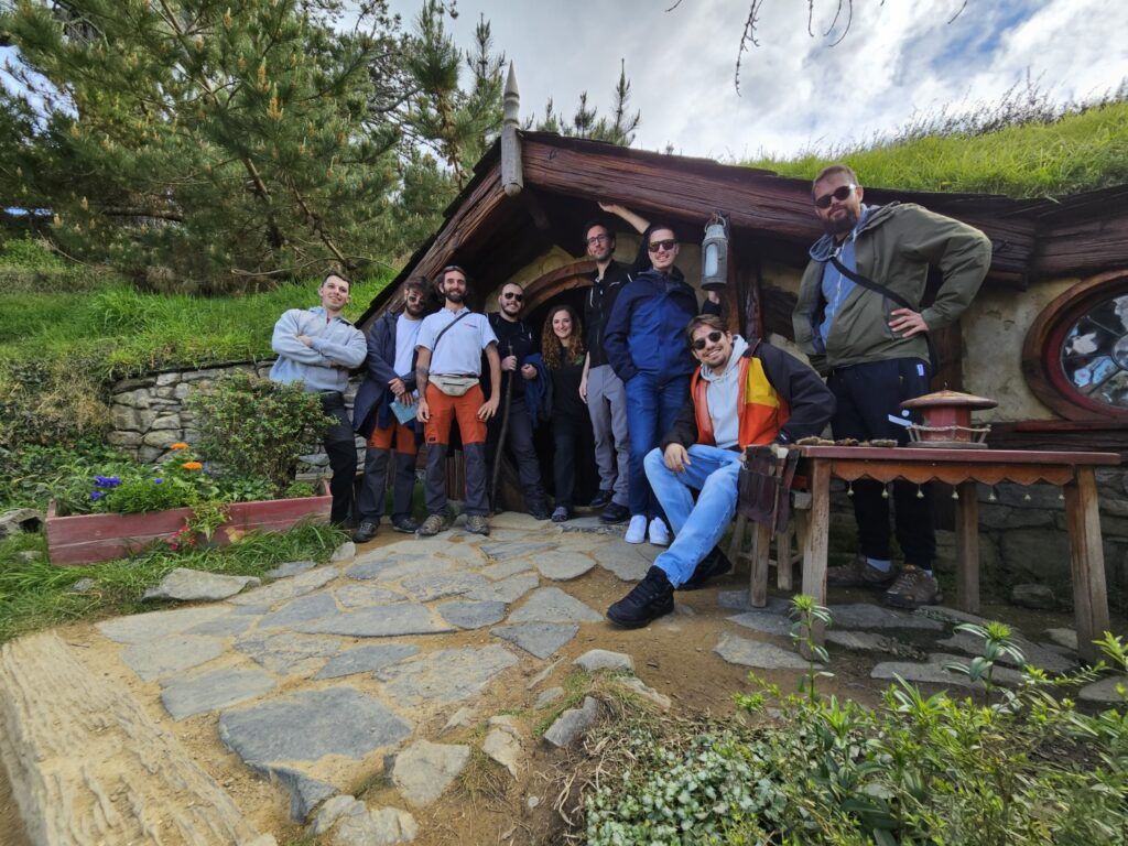 Eine Gruppe von WeRoad-Reisenden posiert vor einem Hobbithaus mit rundem Fenster und Grasdach am Hobbiton-Filmset in Neuseeland.