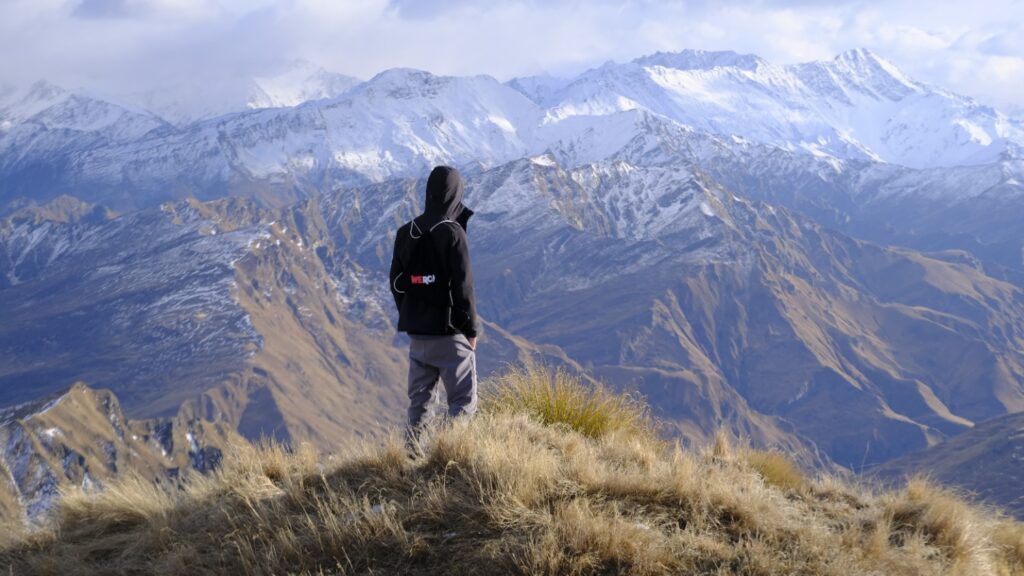 Ein Wanderer steht auf einem grasbewachsenen Gipfel und blickt auf die schneebedeckten Südalpen Neuseelands unter einem bewölkten Himmel.